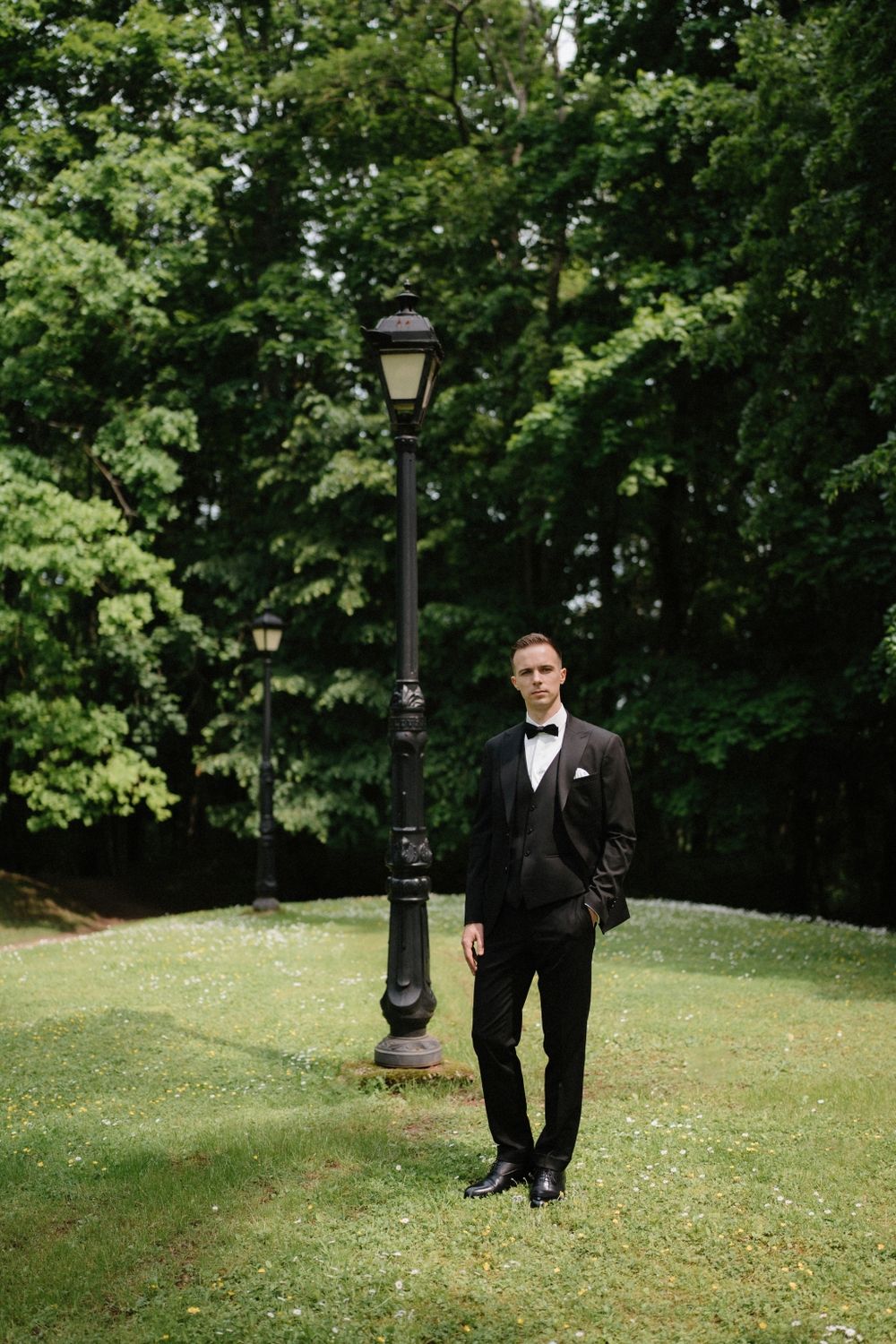 A groom in a classic black tuxedo walks through a park with vintage lampposts.