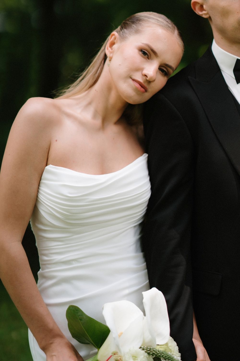 Elegant bride in strapless white dress holds white bouquet while standing next to groom in black suit.