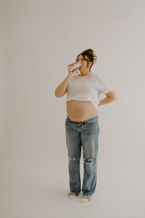 Pregnant woman in casual attire poses against white background while drinking from a bottle.