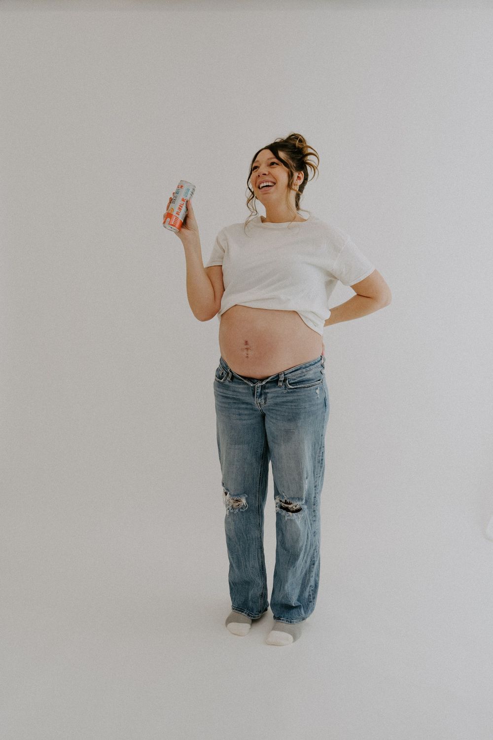 Pregnant woman in distressed jeans holds bottle while posing against white background.