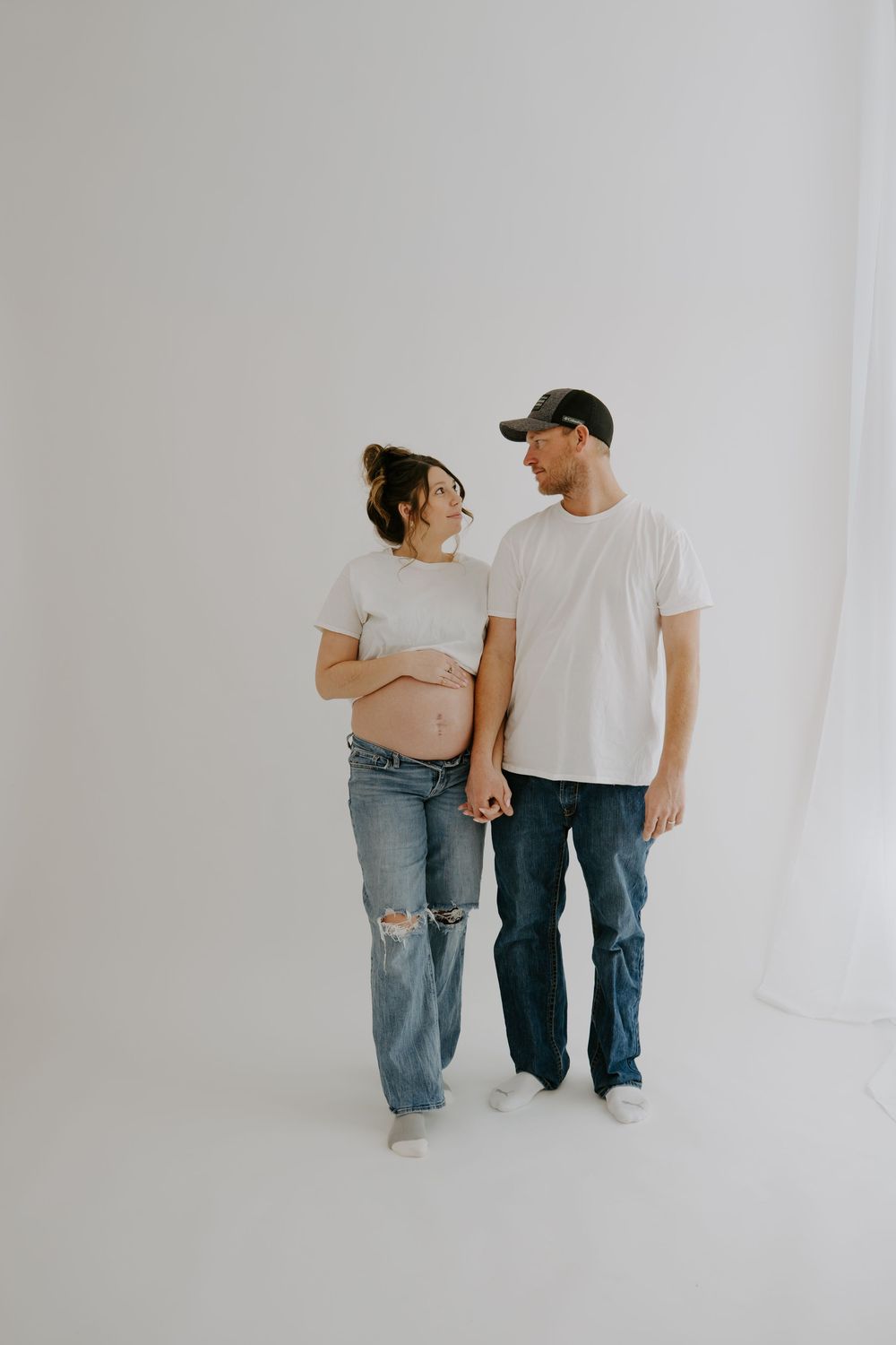 Minimalist maternity photo of couple in white t-shirts and jeans against white backdrop.