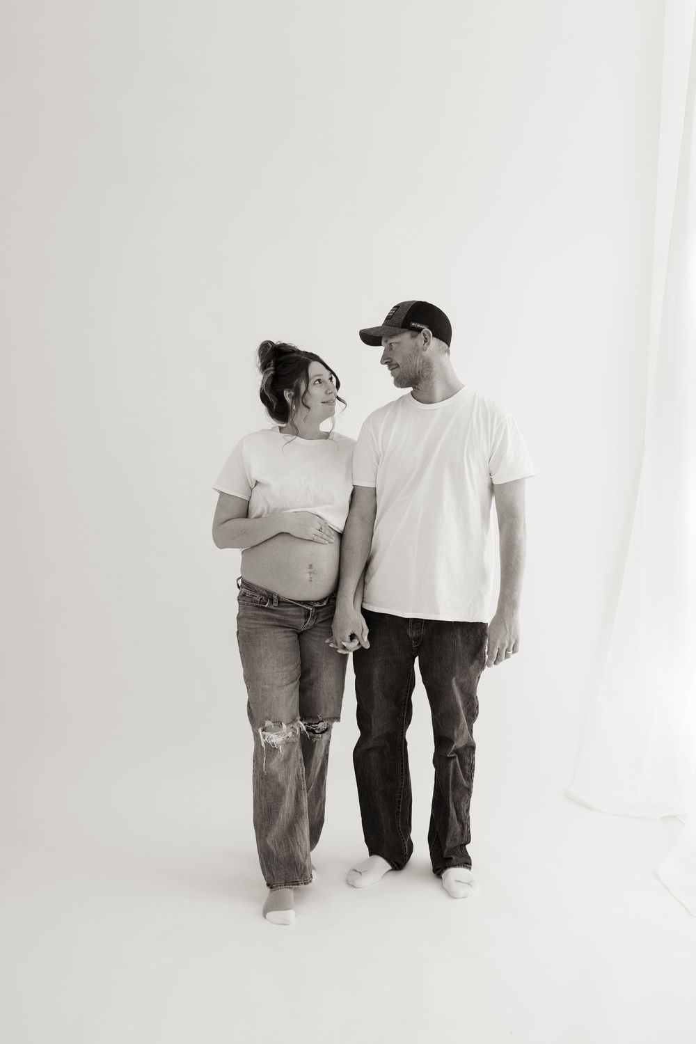 Black and white portrait of expecting couple in casual wear standing against white wall.