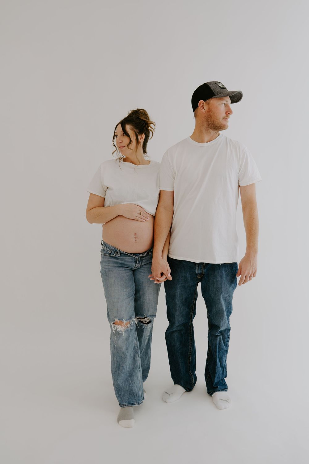 Maternity photo of couple in matching white t-shirts and denim against minimalist background.
