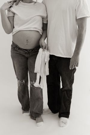Black and white photo of expecting couple with knit baby item against minimal backdrop.