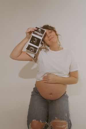 Pregnant woman in ripped jeans holds ultrasound photos against her head while showing her bare belly.