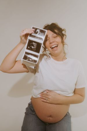 Joyful pregnant woman laughs while holding ultrasound strip photos above her exposed belly.