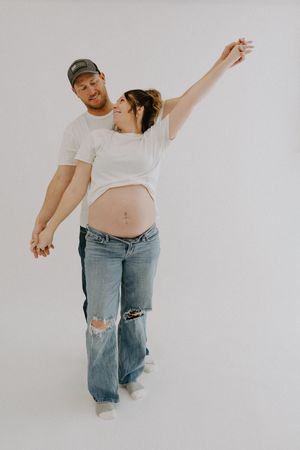 Expectant couple poses playfully in casual attire against white backdrop.