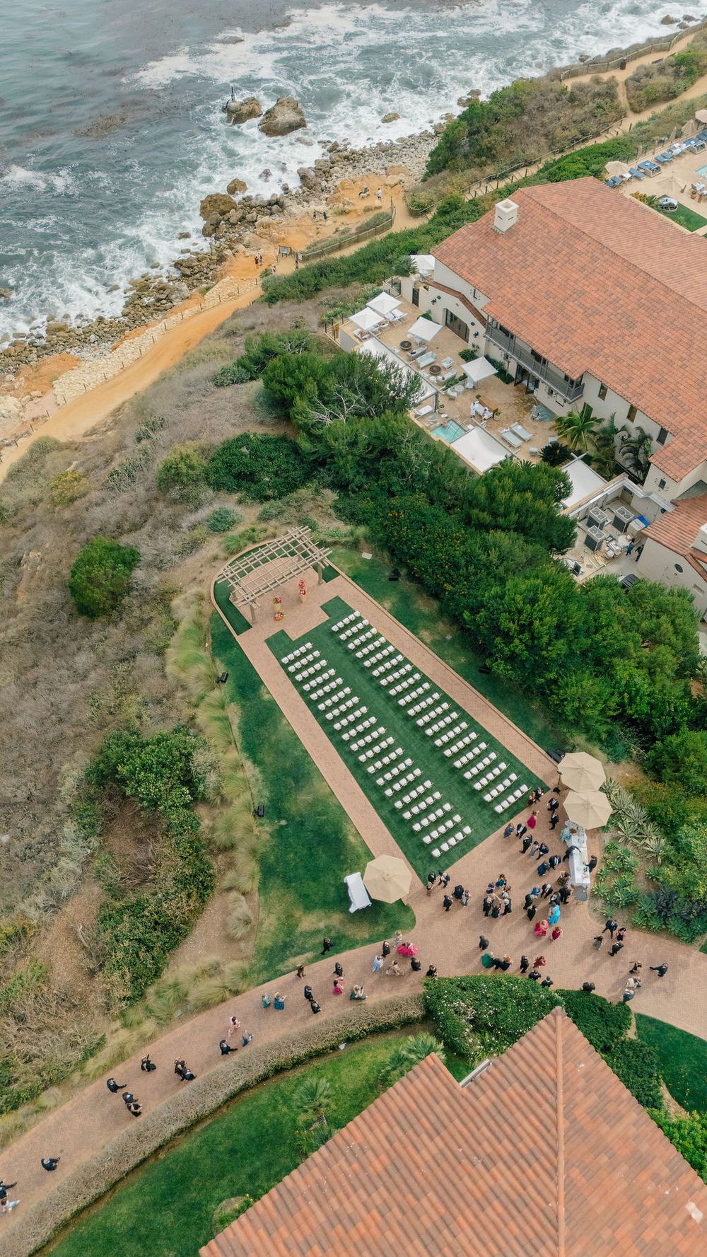 Aerial view of an outdoor wedding ceremony setup on a coastal cliff with rows of white chairs and ocean views.
