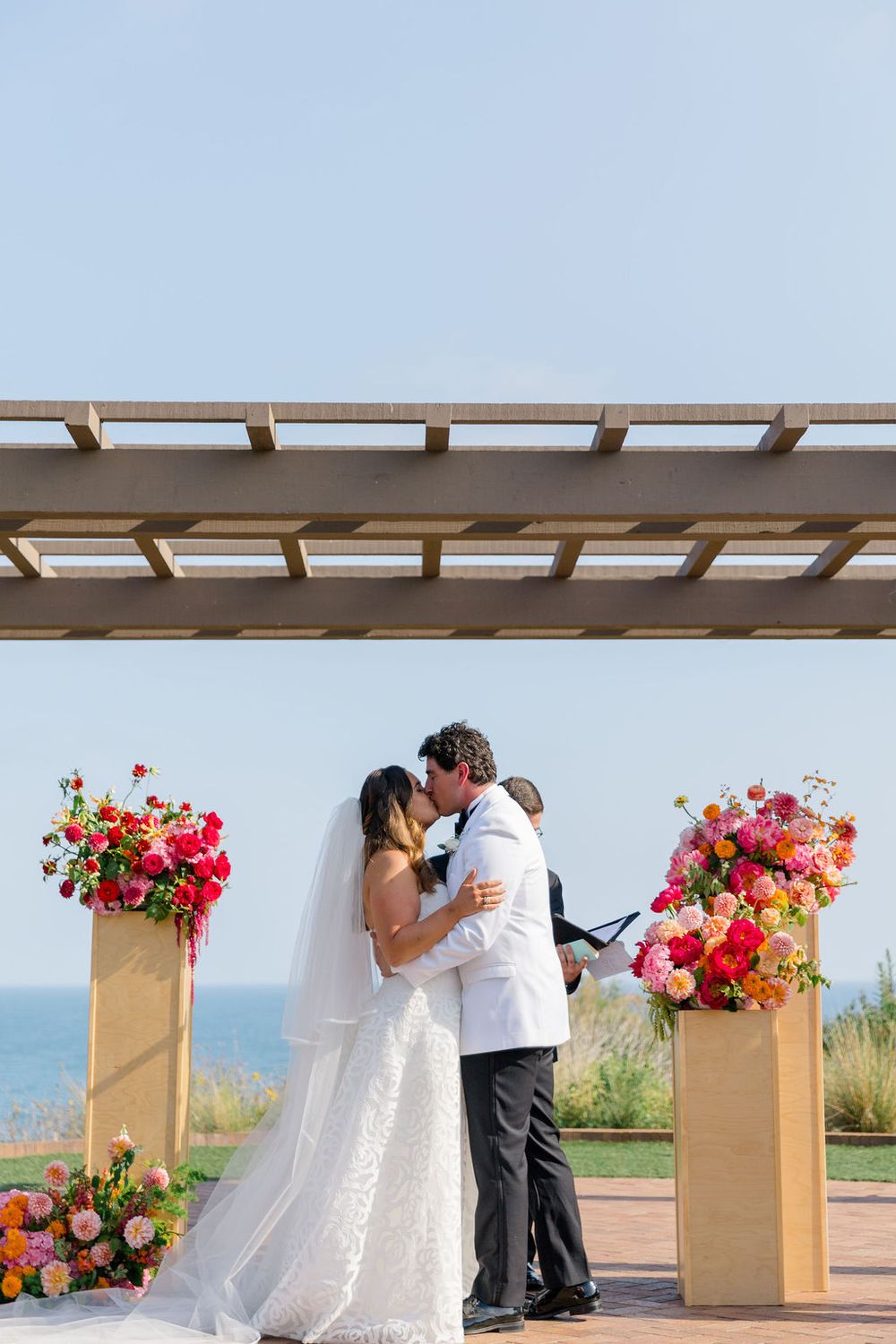 A romantic outdoor wedding ceremony on a coastal terrace with vibrant coral and pink floral arrangements.