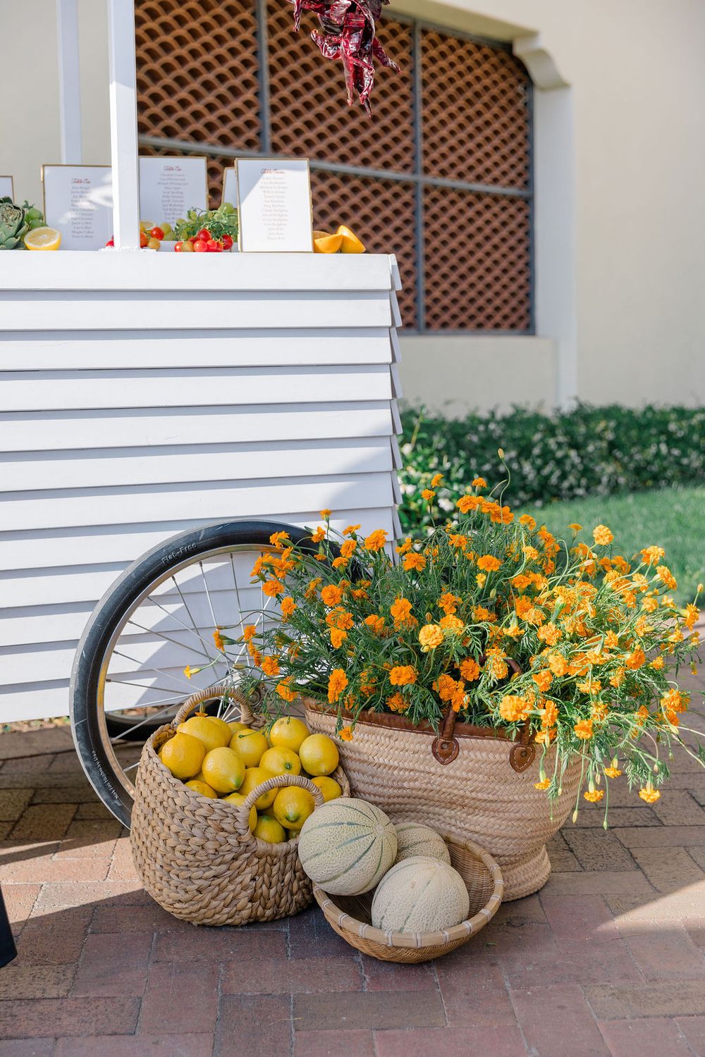 White cart display with vibrant marigolds and citrus fruits in woven baskets on brick patio.
