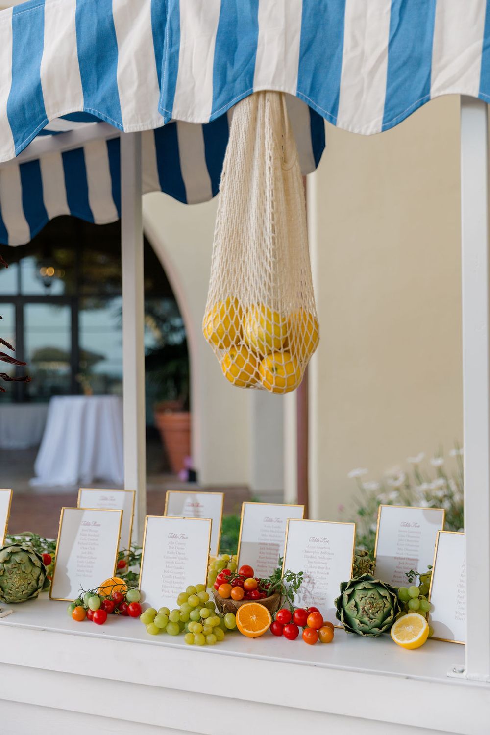 Fresh fruit and menu cards displayed under a blue and white striped awning with hanging lemons in mesh netting.