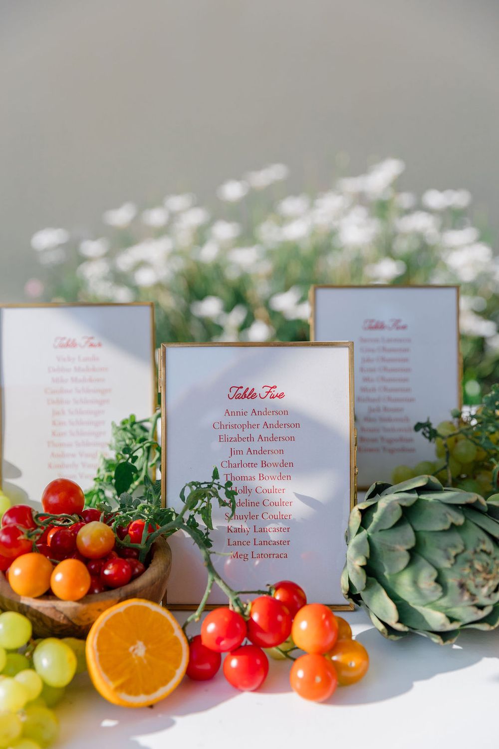 Wedding table cards displayed with fresh citrus fruits, tomatoes, artichoke and greenery on white surface.