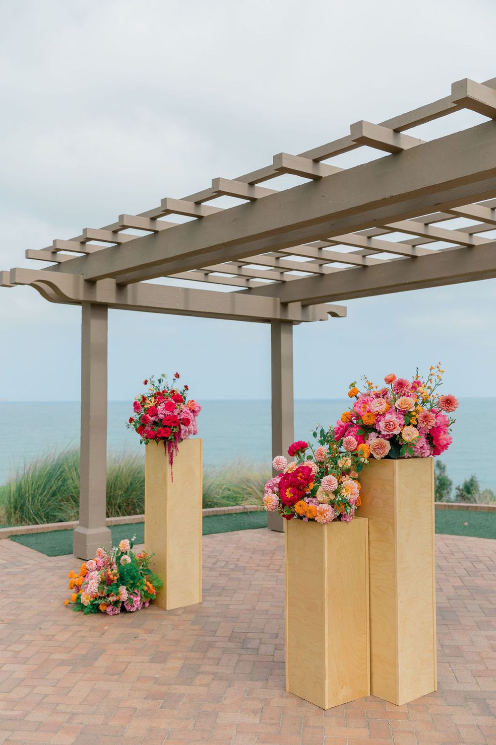 Beige wooden pergola with gold pedestals and vibrant pink and red floral arrangements on a tiled patio overlooking a scenic view.