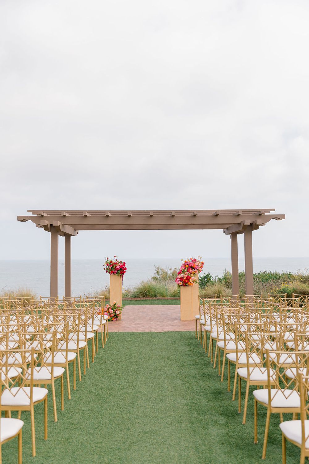 Outdoor wedding ceremony setup with wooden chairs and pergola overlooking the ocean on a grassy lawn.