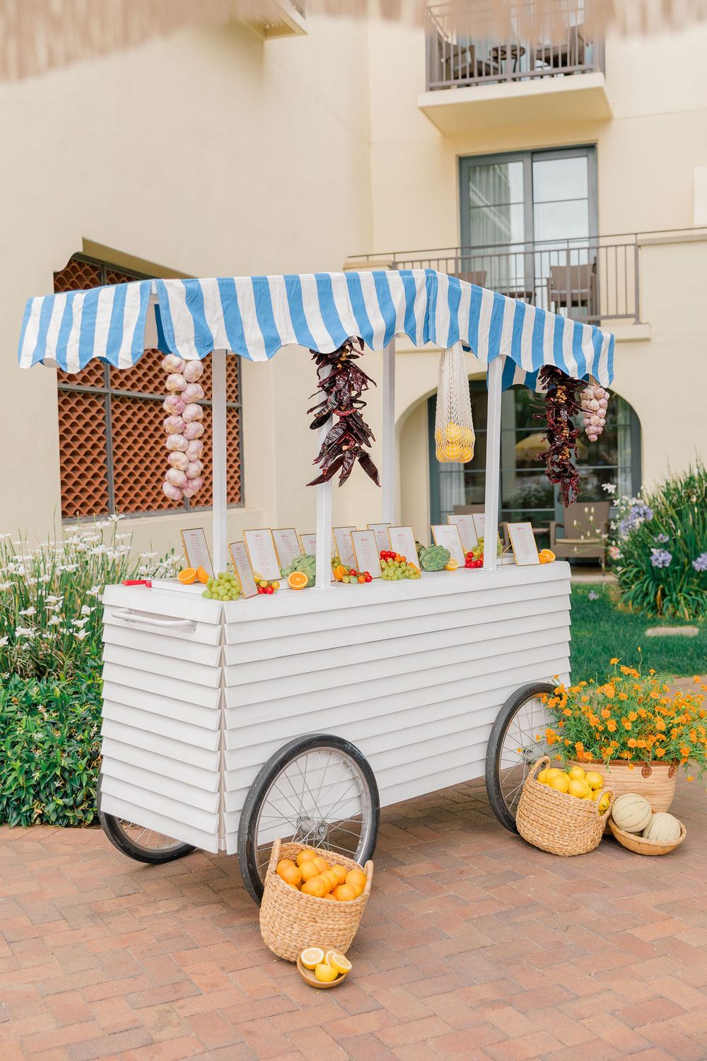 A charming white vendor cart with blue and white striped awning displays fresh produce and flowers on a terracotta patio.