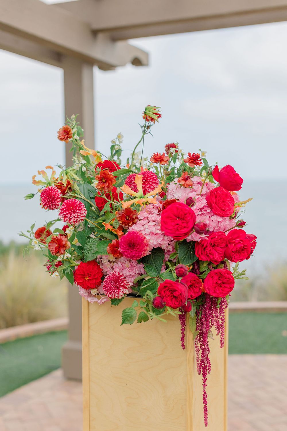 Red roses and pink dahlias arranged in modern blonde wood pedestals against a coastal backdrop.
