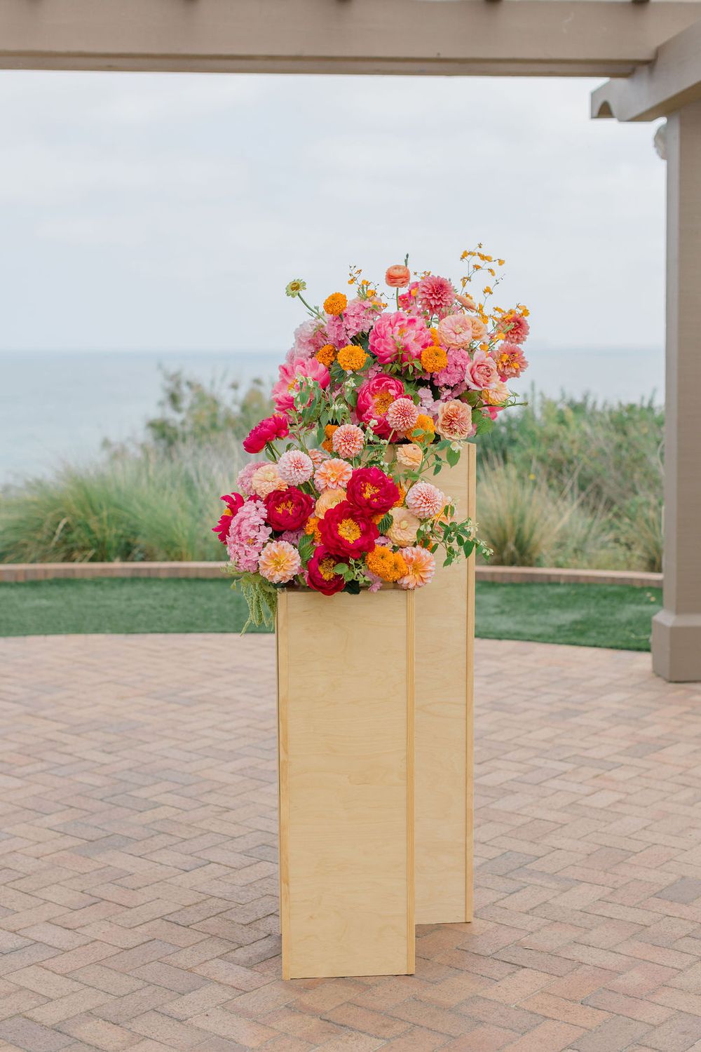 Three wooden planter boxes filled with colorful summer flowers on a herringbone brick patio overlooking the ocean.