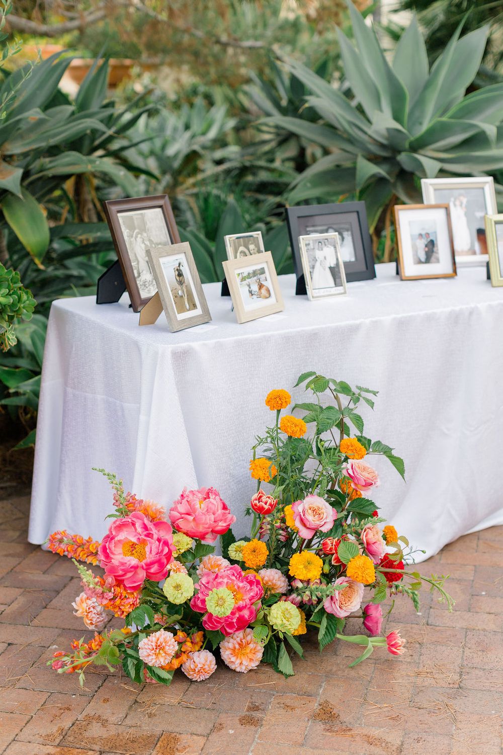 White tables with framed photographs and floral arrangements in coral and orange hues displayed against lush garden backdrop.