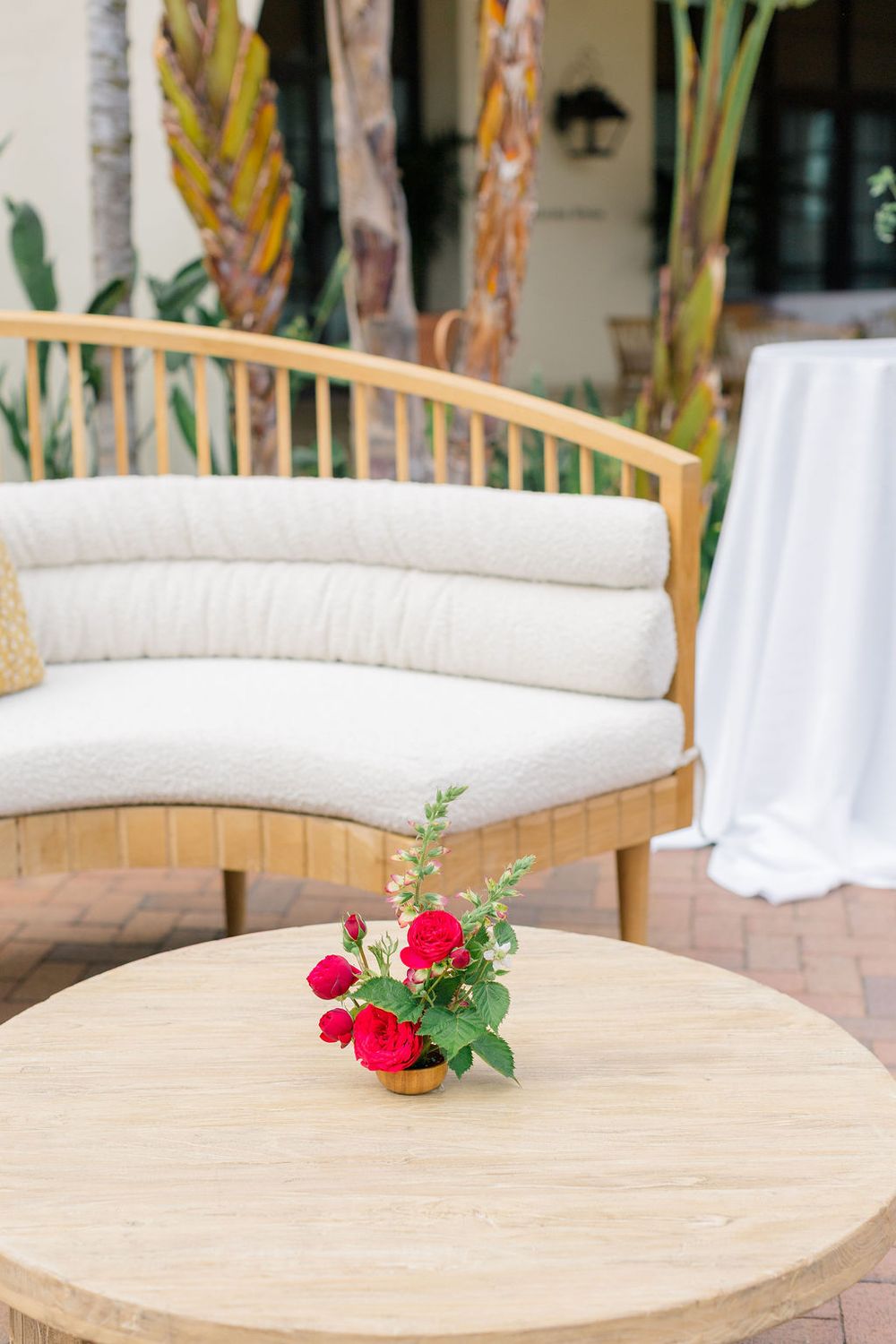 Curved white bench seating with red floral centerpiece on light wood table at outdoor venue.