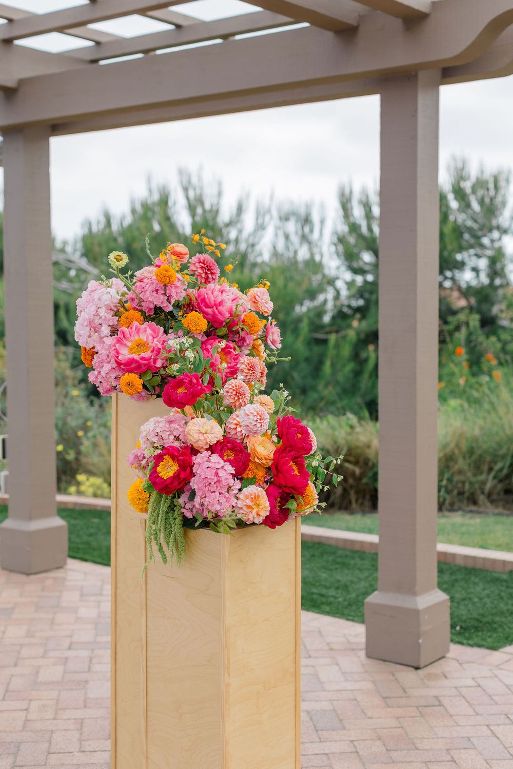 Vibrant pink and orange floral arrangements in wooden boxes line an outdoor covered patio with stone flooring.