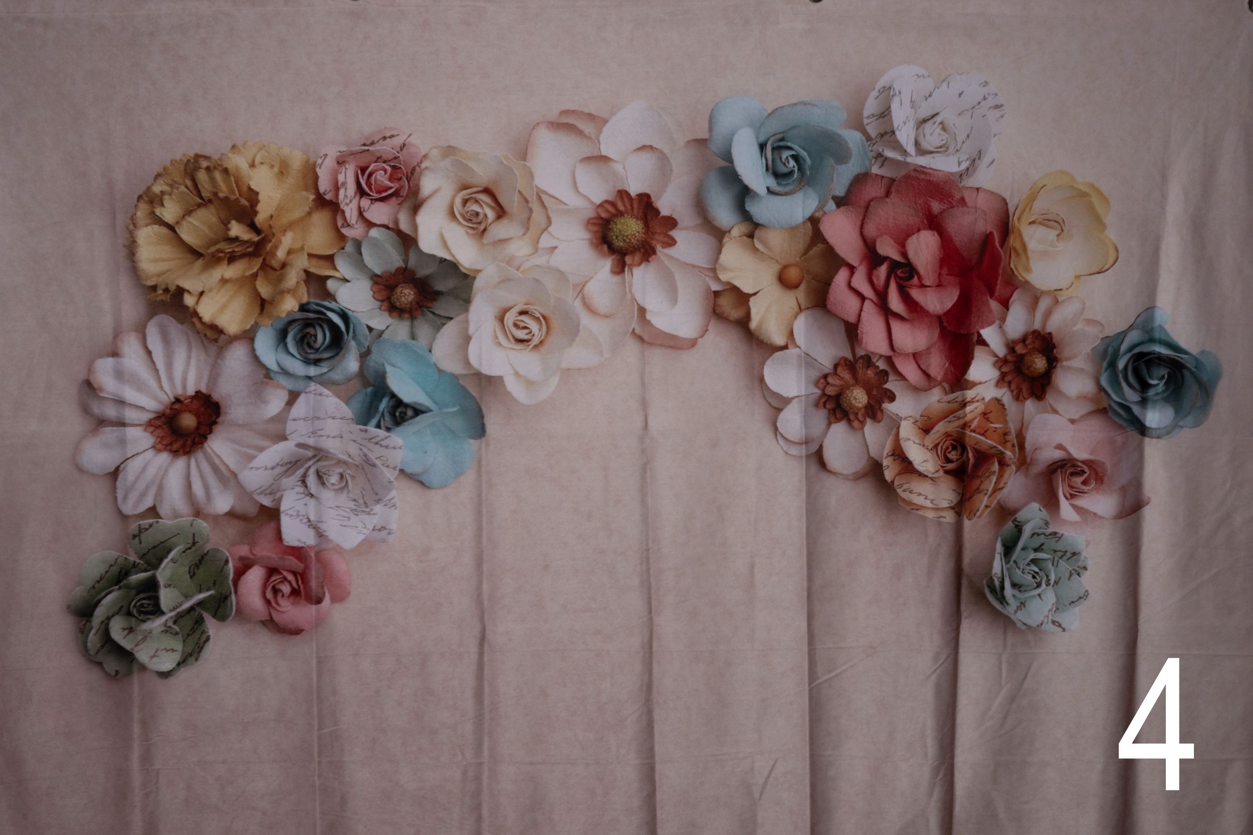 Delicate paper flowers in soft pastel colors of pink, blue, and cream arranged along a textured gray wall.