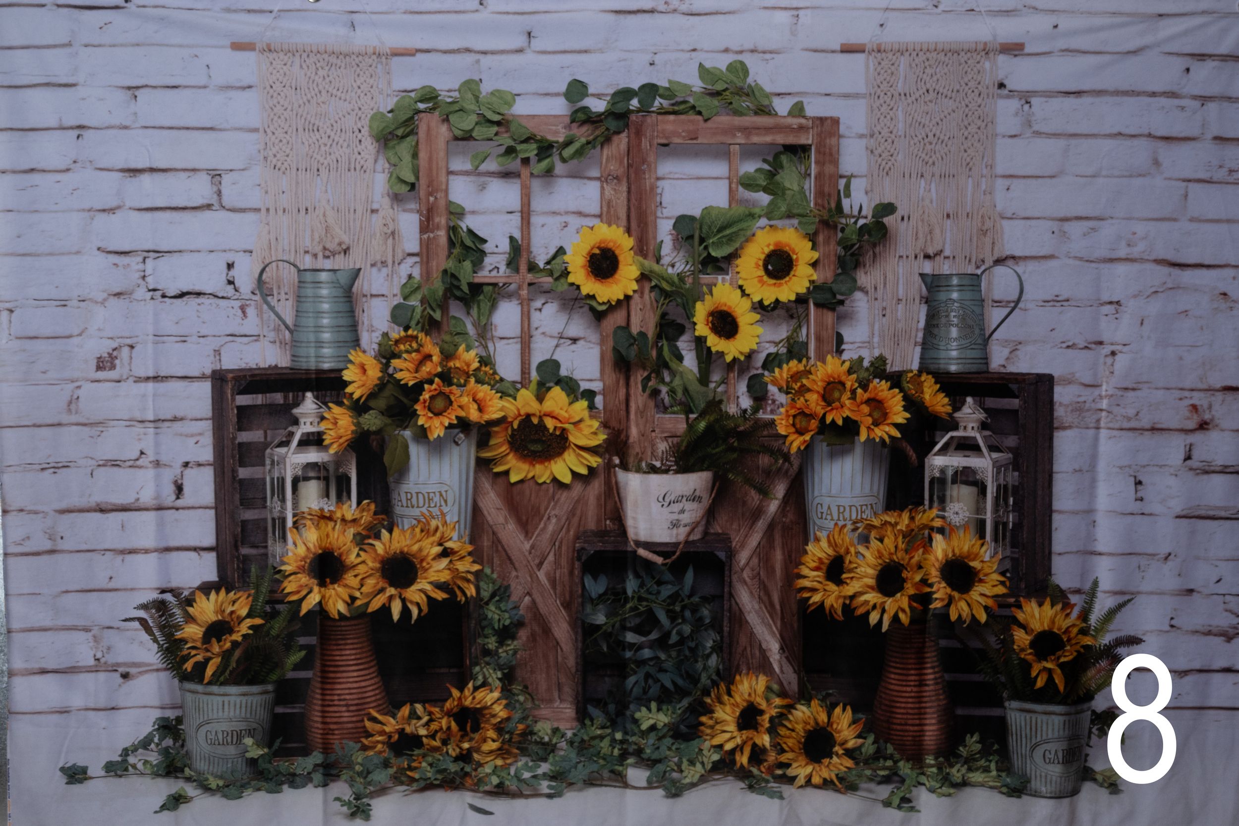 Rustic white brick wall decorated with sunflower garlands, wooden frames, and potted plants creates a charming farmhouse display.