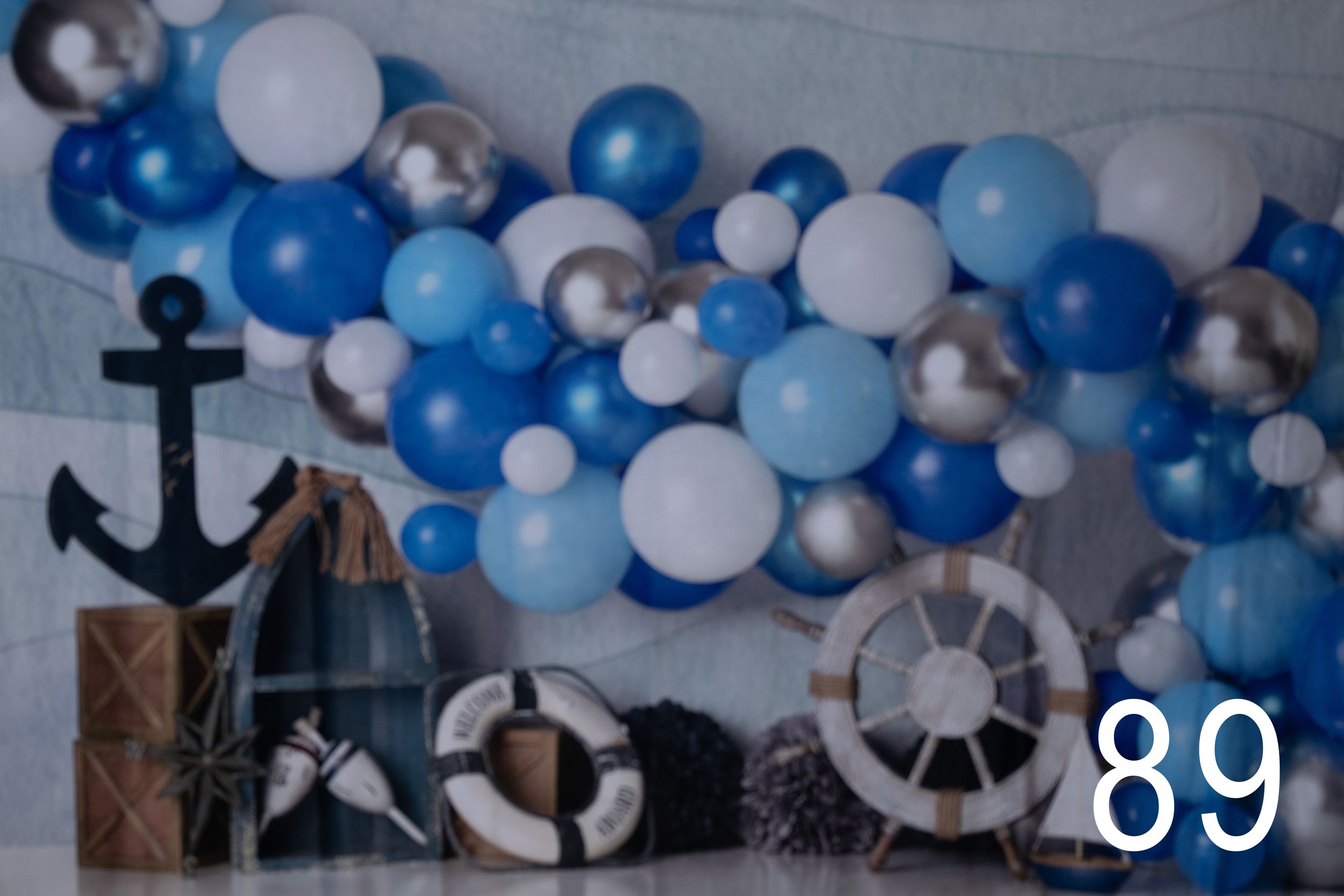 Nautical themed party decoration with blue and white balloon garland, anchor, and ship wheel against gray wall.