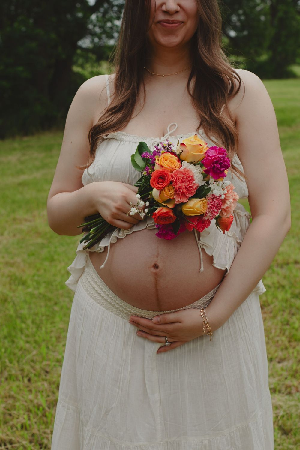 Maternity photo showing colorful flowers held against a pregnant belly in a white dress outdoors.