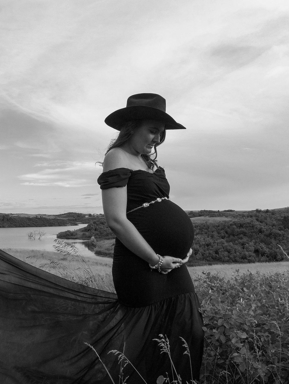 Dramatic black and white maternity silhouette against cloudy sky wearing flowing dress.
