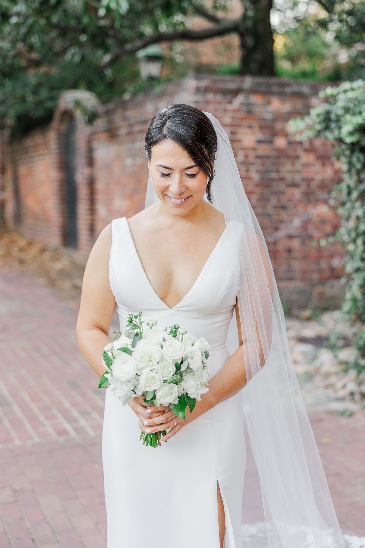 A bride in a white V-neck wedding dress holds a classic white and green bouquet against a brick wall backdrop.