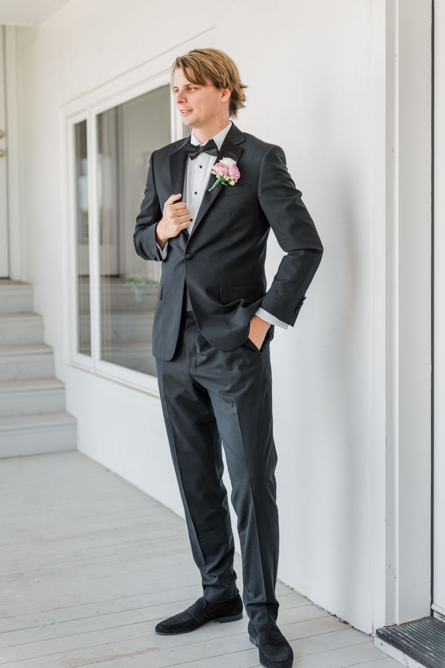 Groom in classic black tuxedo poses confidently against white wall during wedding preparations.