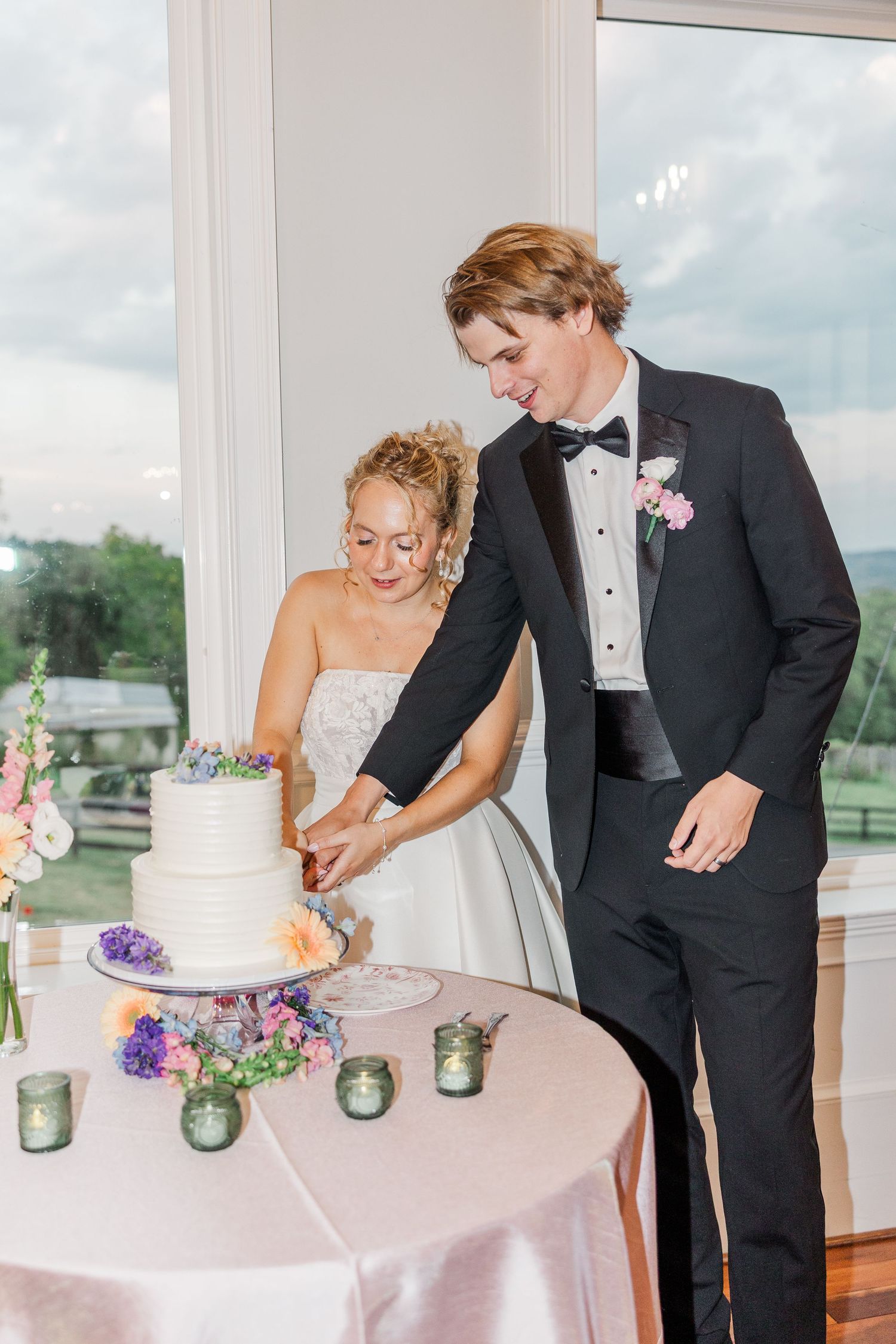Couple cuts their white wedding cake decorated with purple flowers at their reception.