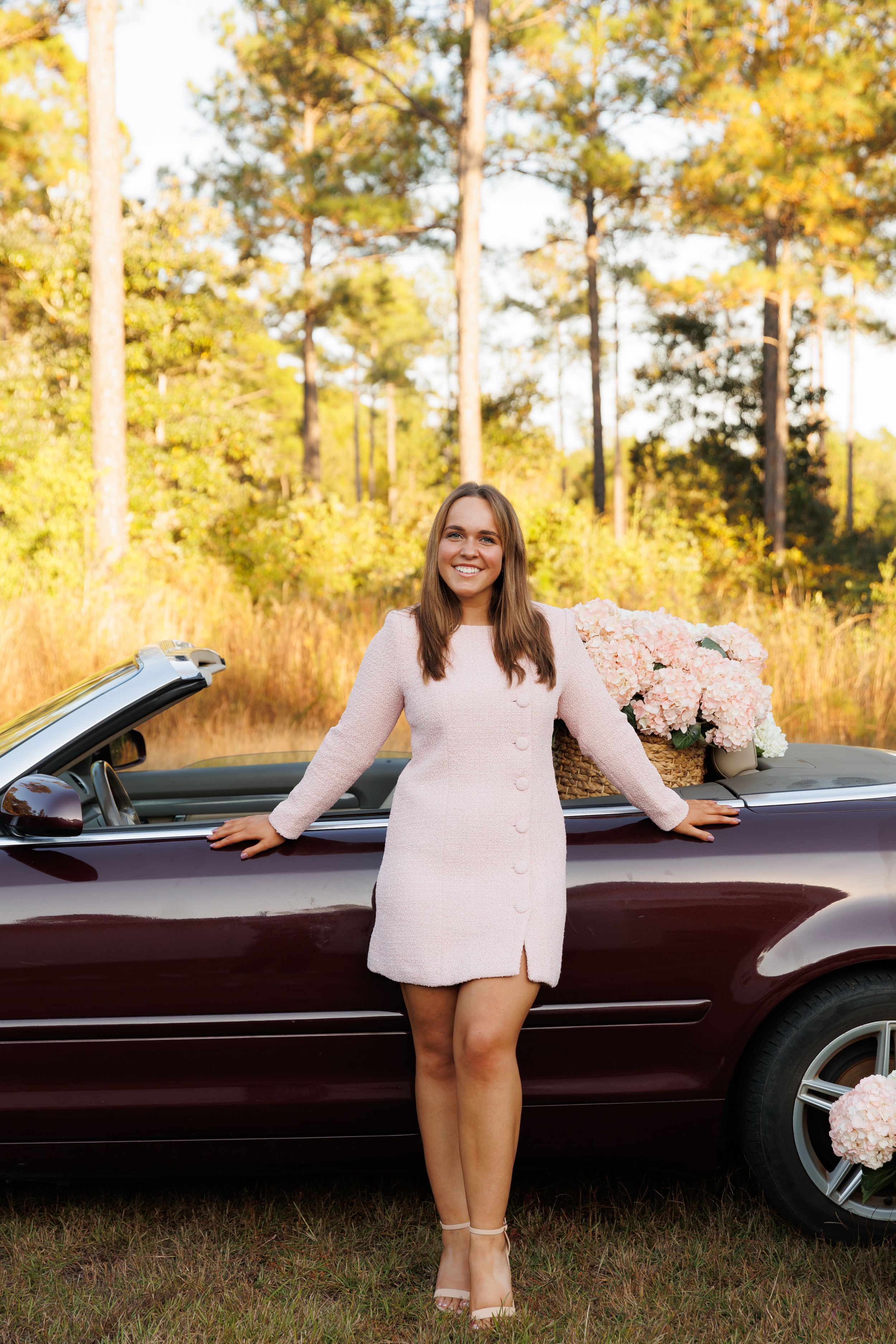 A pink-dressed figure poses beside a burgundy convertible car in a sunlit wooded setting with golden fall foliage.