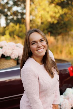 A person wearing a soft pink sweater smiles warmly while sitting outdoors in autumn with golden foliage in the background.