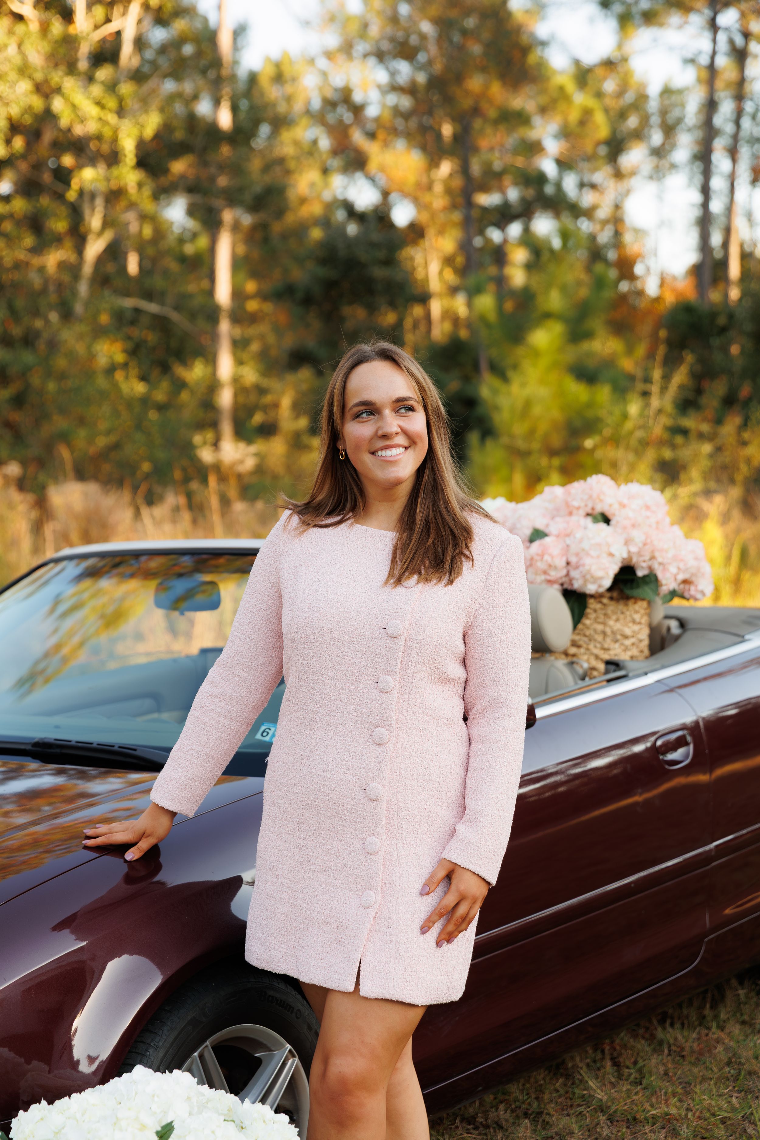 A person in light pink attire stands next to a dark burgundy luxury car against a backdrop of autumn trees.