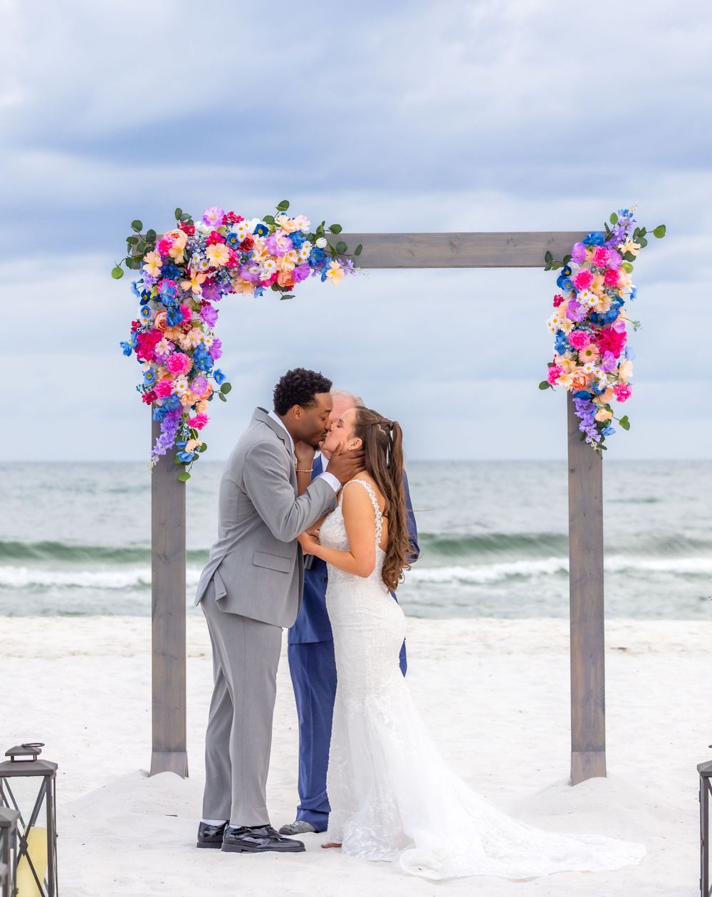 A couple shares a romantic moment under a floral arch on a cloudy beach with waves in the background.