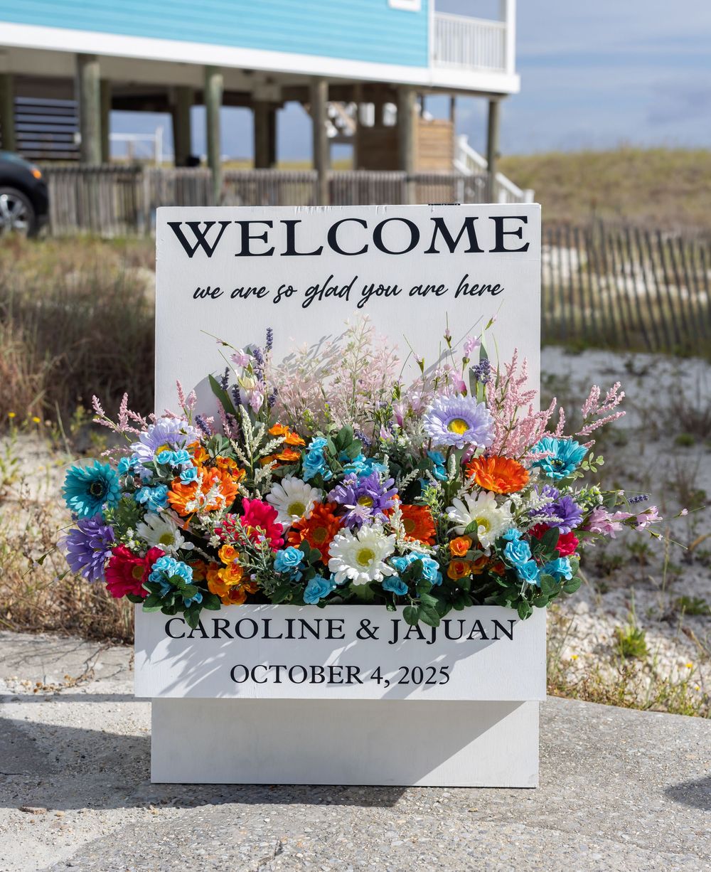 Wedding welcome sign with colorful floral arrangements displayed on a sandy beach with ocean waves in the background.