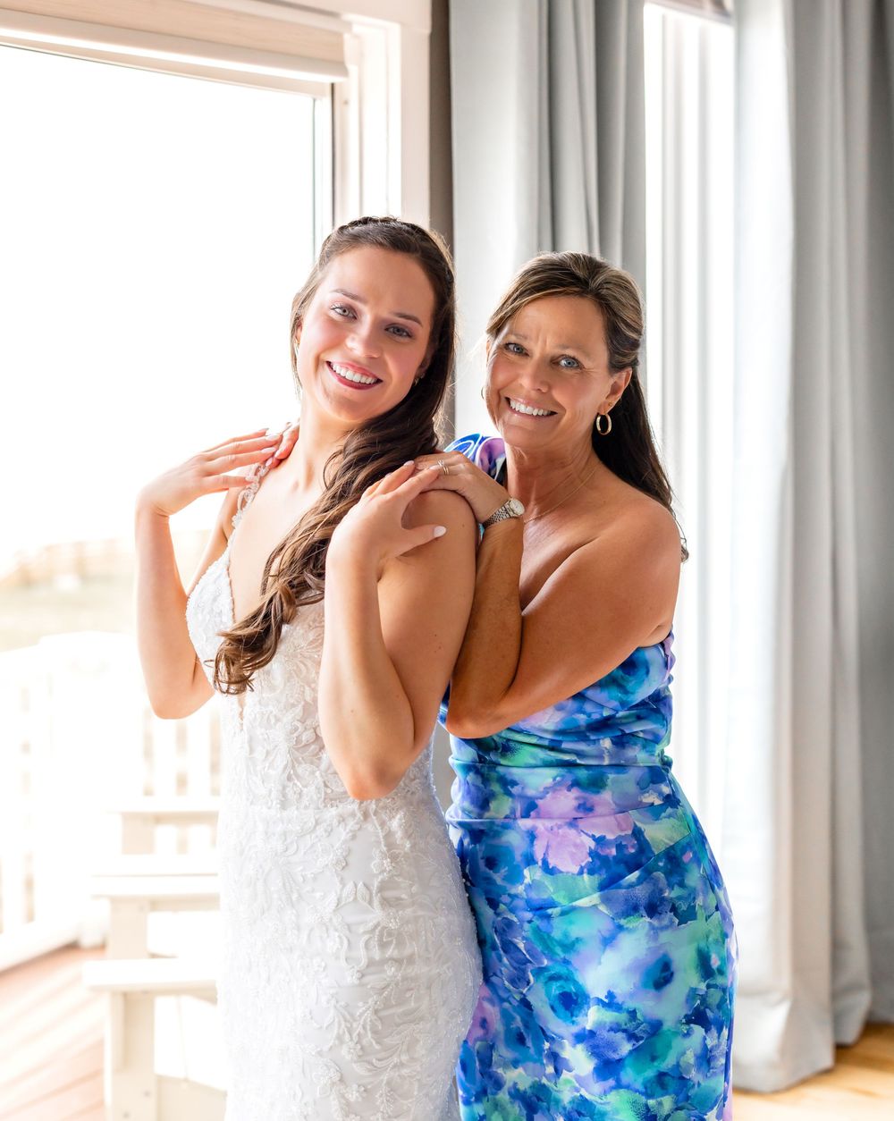 Two people laughing and embracing while wearing white and blue floral dresses in a bright indoor setting.