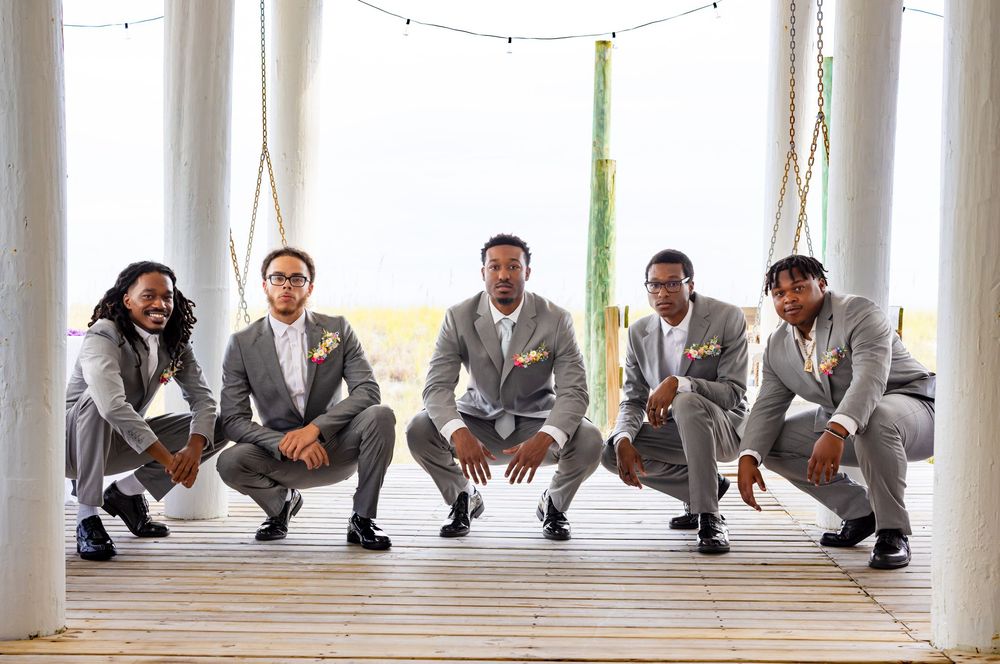 A group of groomsmen in matching gray suits crouch together in a bright white room with natural lighting.