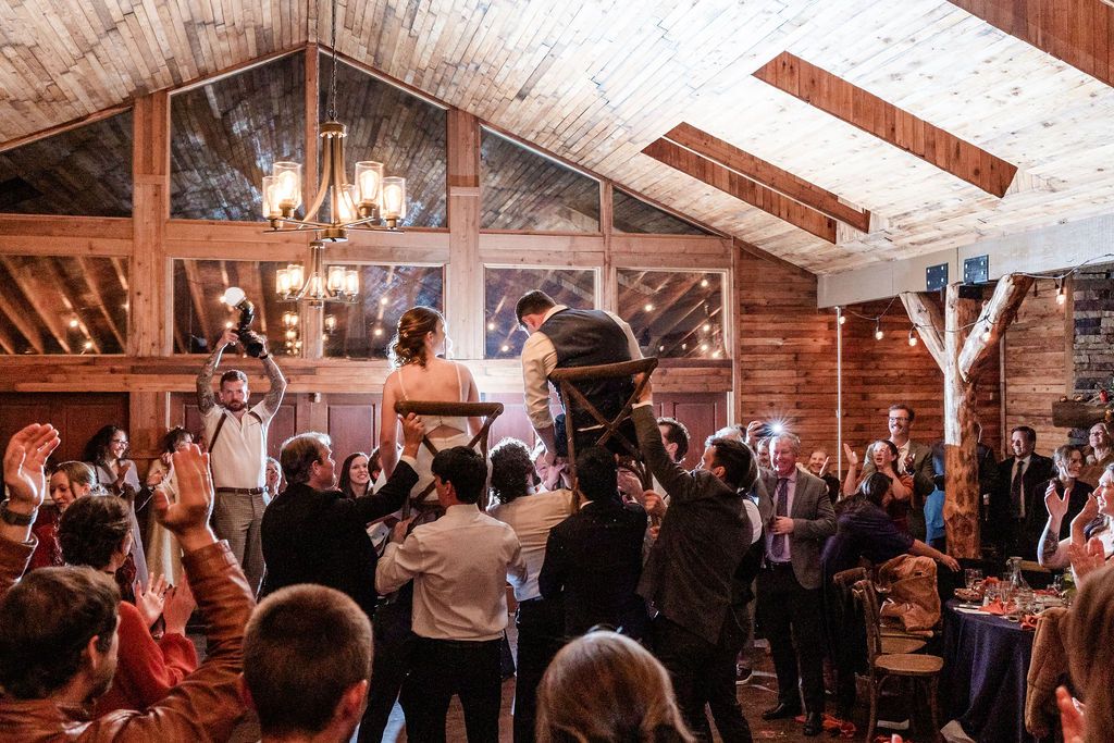 Wedding guests dance and celebrate in a rustic wooden barn venue with exposed beams and warm lighting.