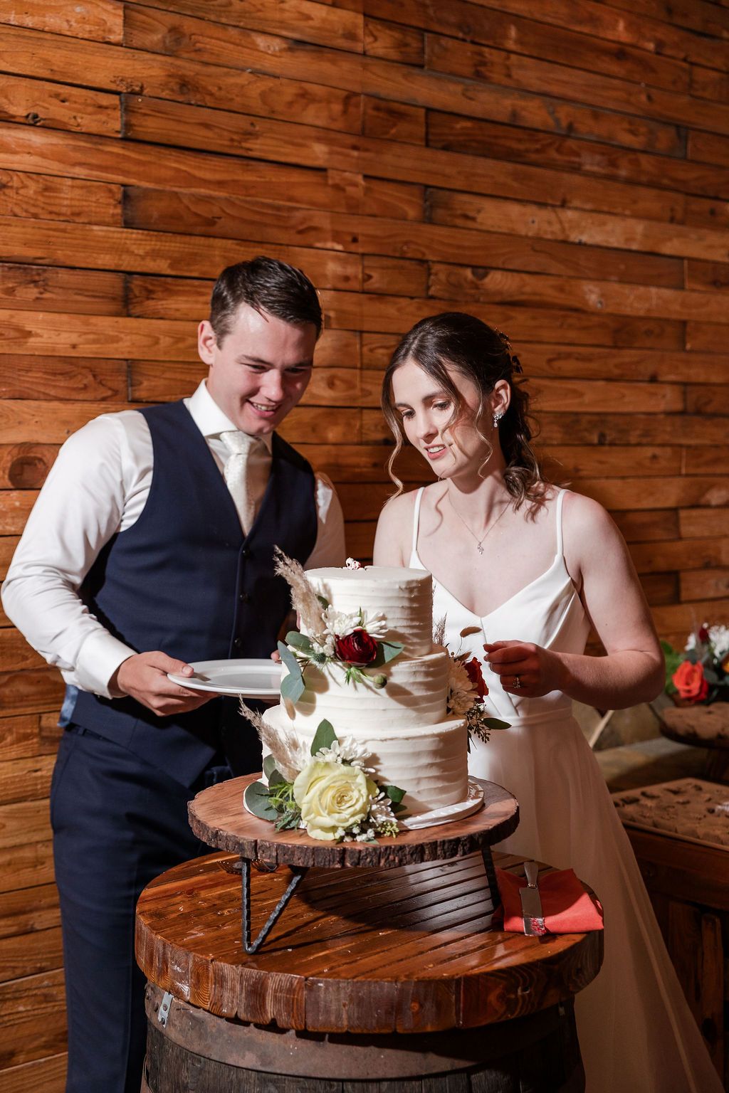 Newlyweds cut into their elegant white tiered wedding cake displayed on rustic wooden barrels against wood paneled wall.