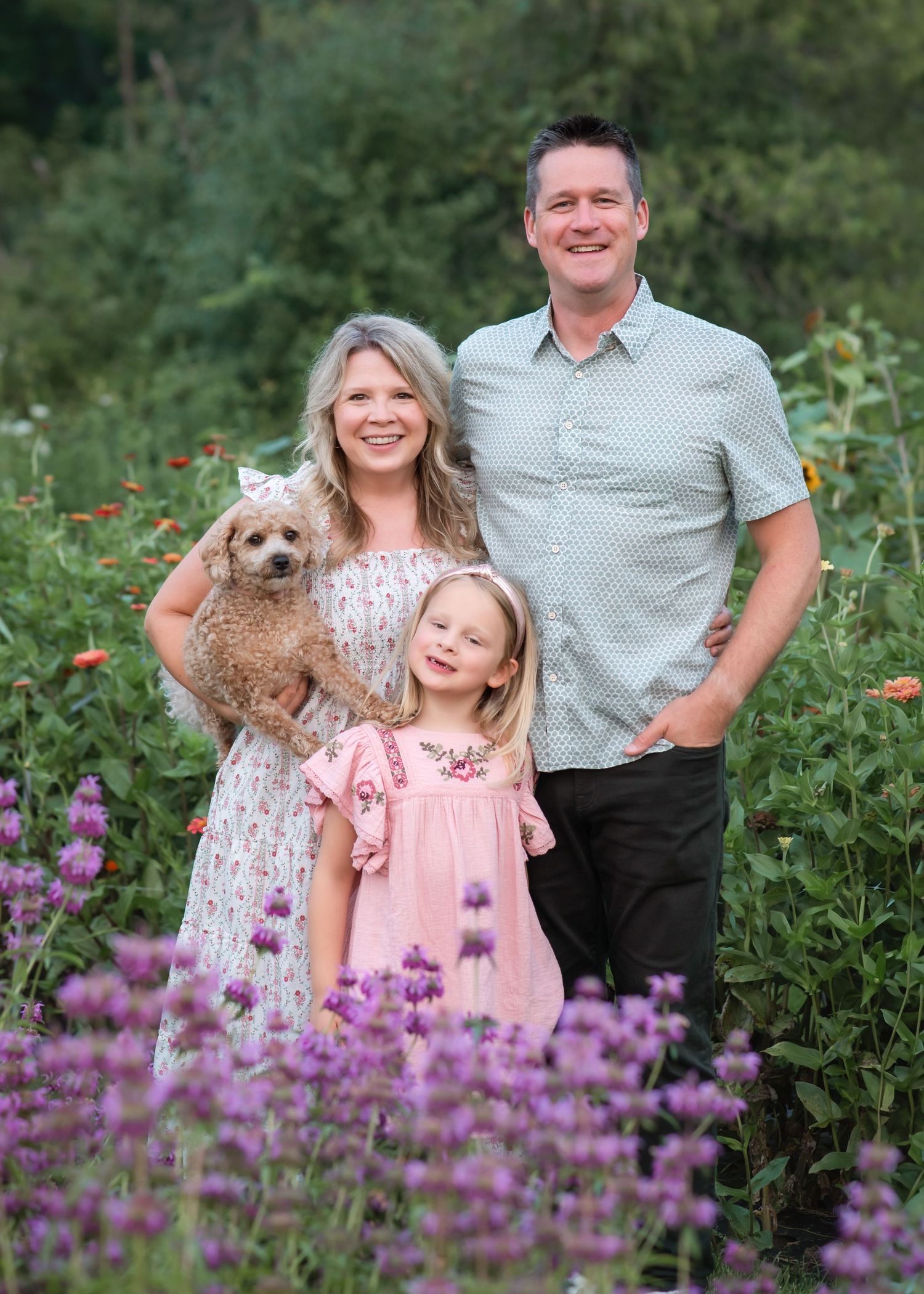 A family poses together in a garden with purple wildflowers and a small goldendoodle dog.