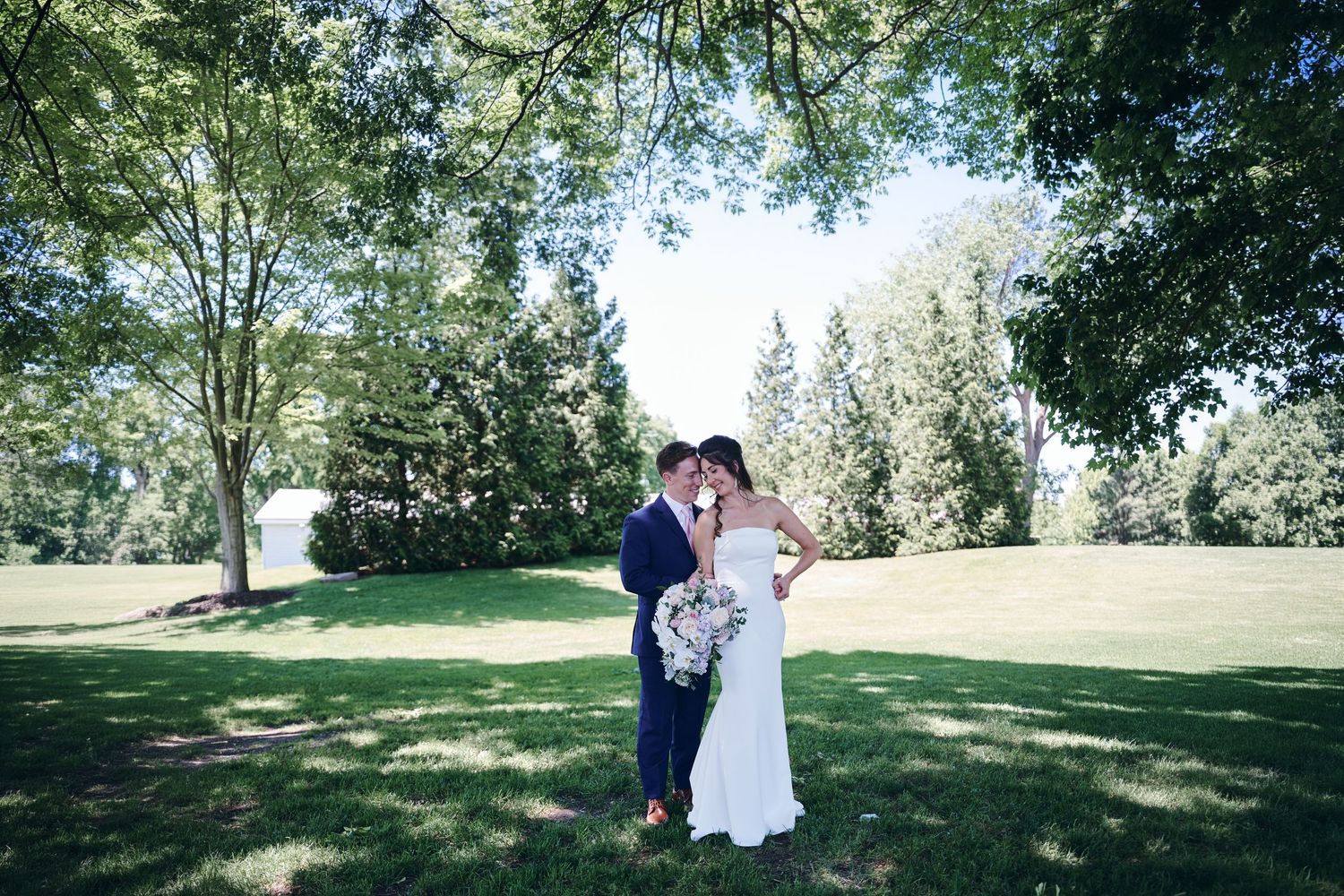 Bride and groom share a romantic kiss outdoors under lush green trees on a sunny wedding day.