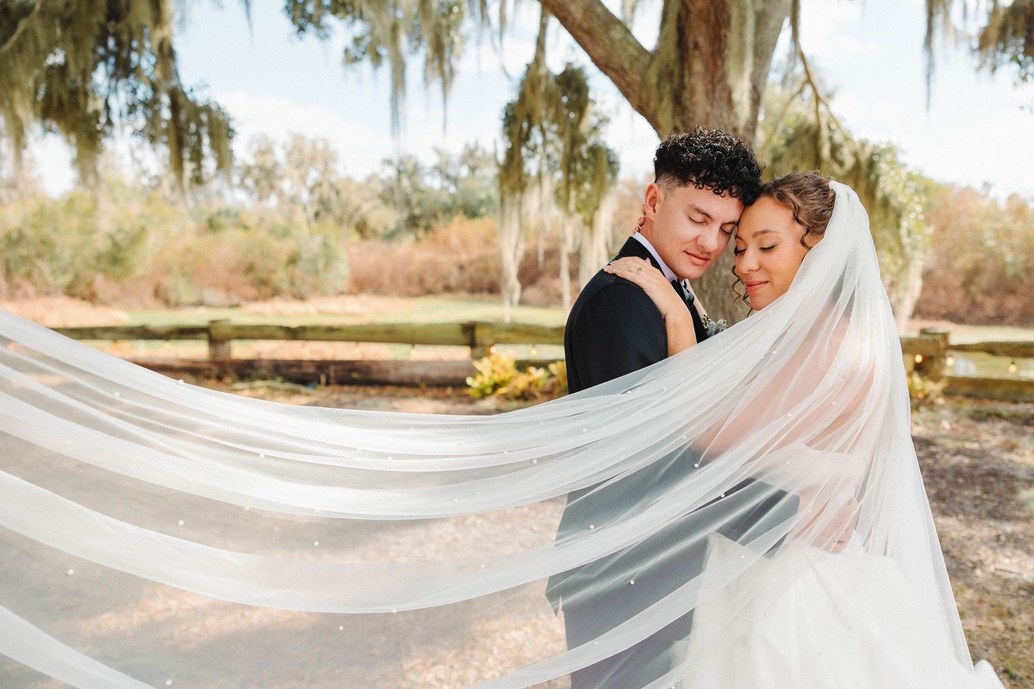 Bride and groom embrace under Spanish moss trees, her flowing veil billowing around them at an outdoor wedding venue.