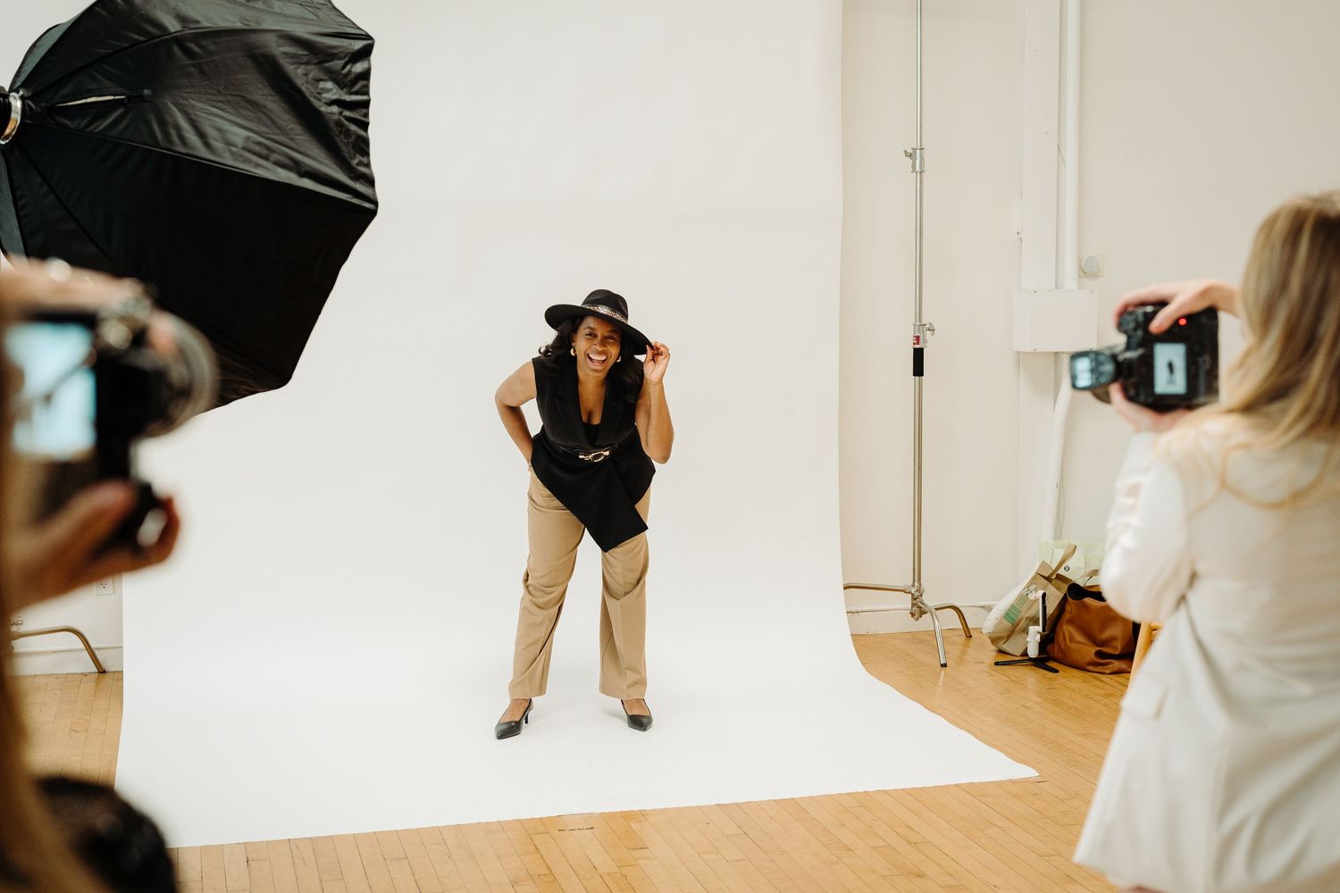 A model poses in a photography studio with a white backdrop, wearing a black top, tan pants, and a hat.