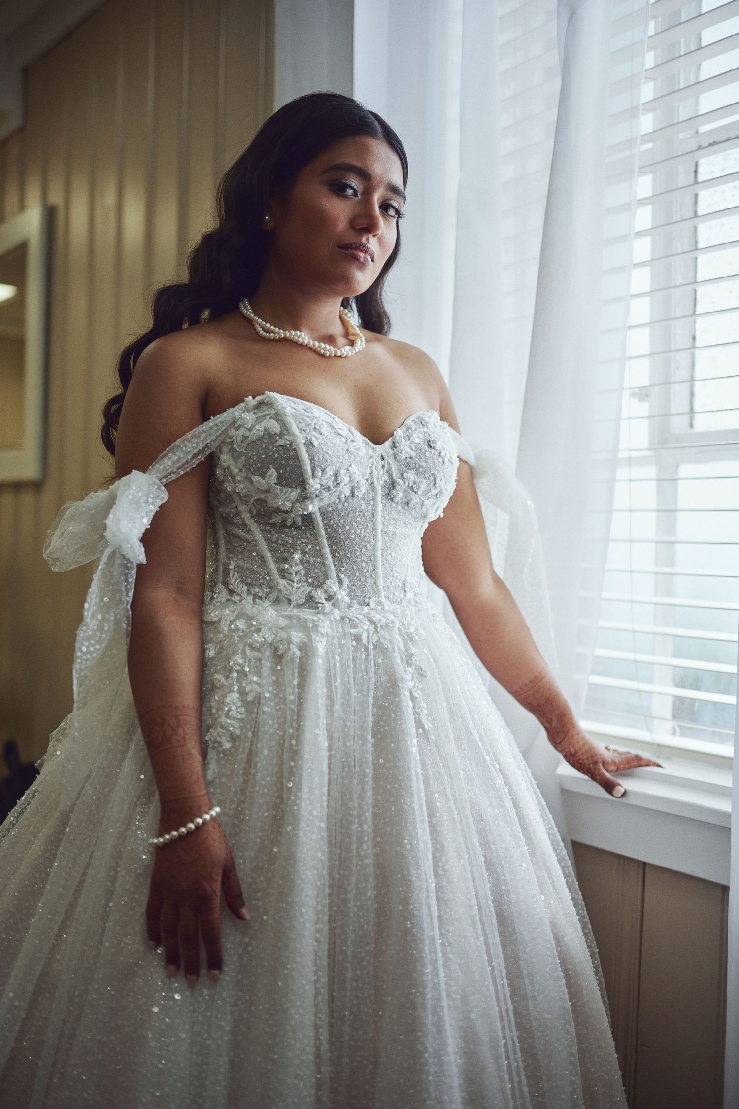 Bride in off-shoulder sparkly white ballgown with lace appliques and pearl necklace poses by window in soft light.