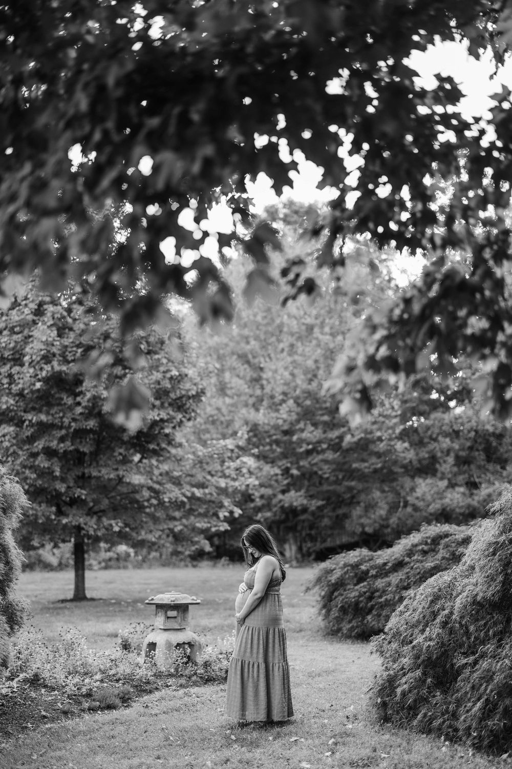 Black and white photo of a pregnant woman in a long dress cradling her belly in a lush garden setting.