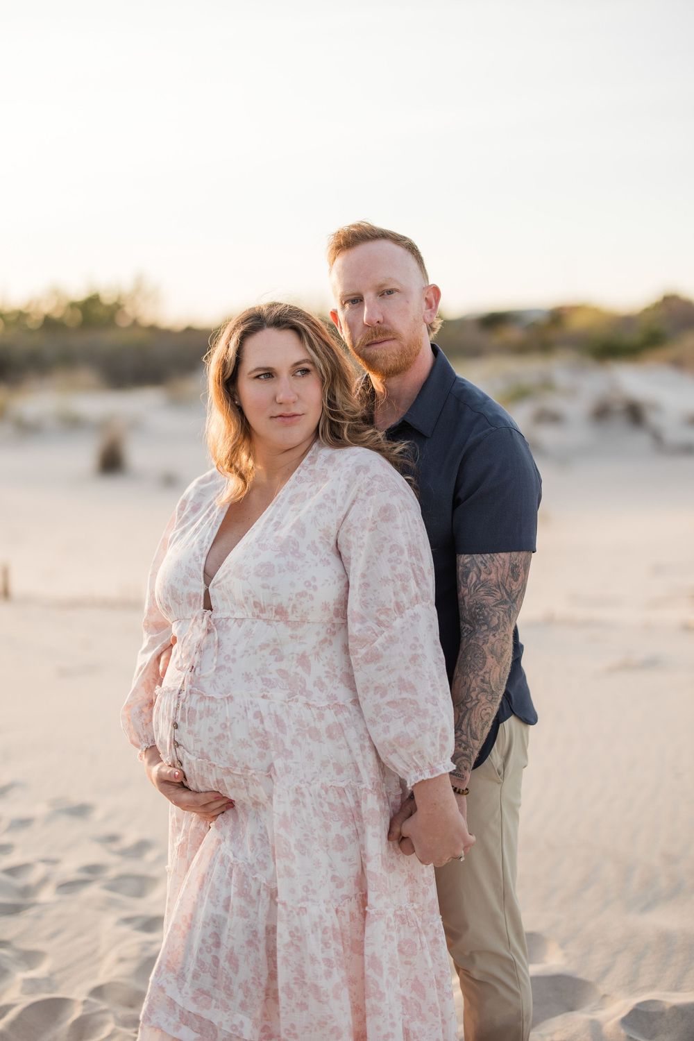 Pregnant couple posing on sandy beach at golden hour, woman in floral dress cradling bump, man with tattoos behind her.