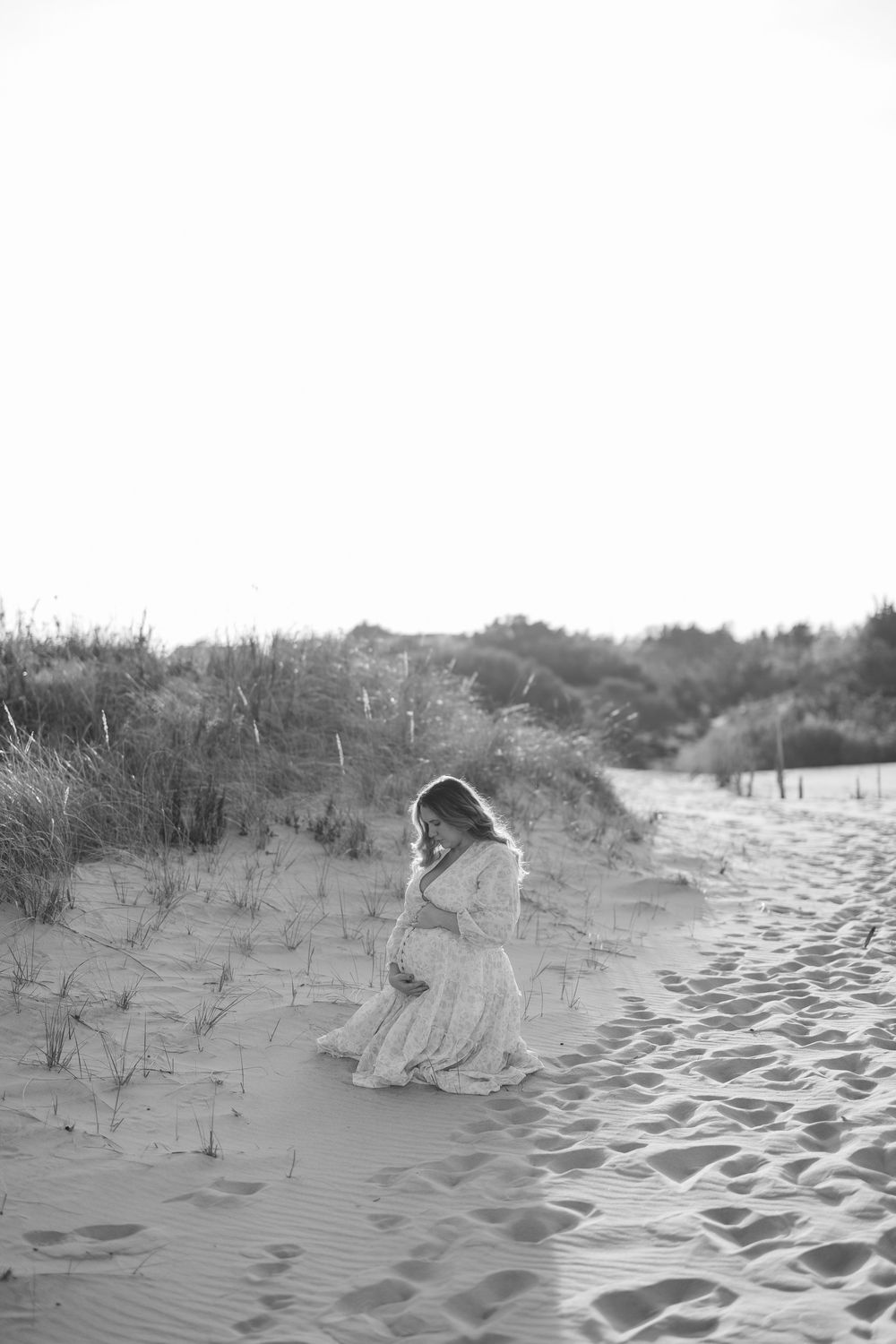 Black and white photo of a pregnant woman in a flowing dress kneeling on sandy dunes, cradling her baby bump.