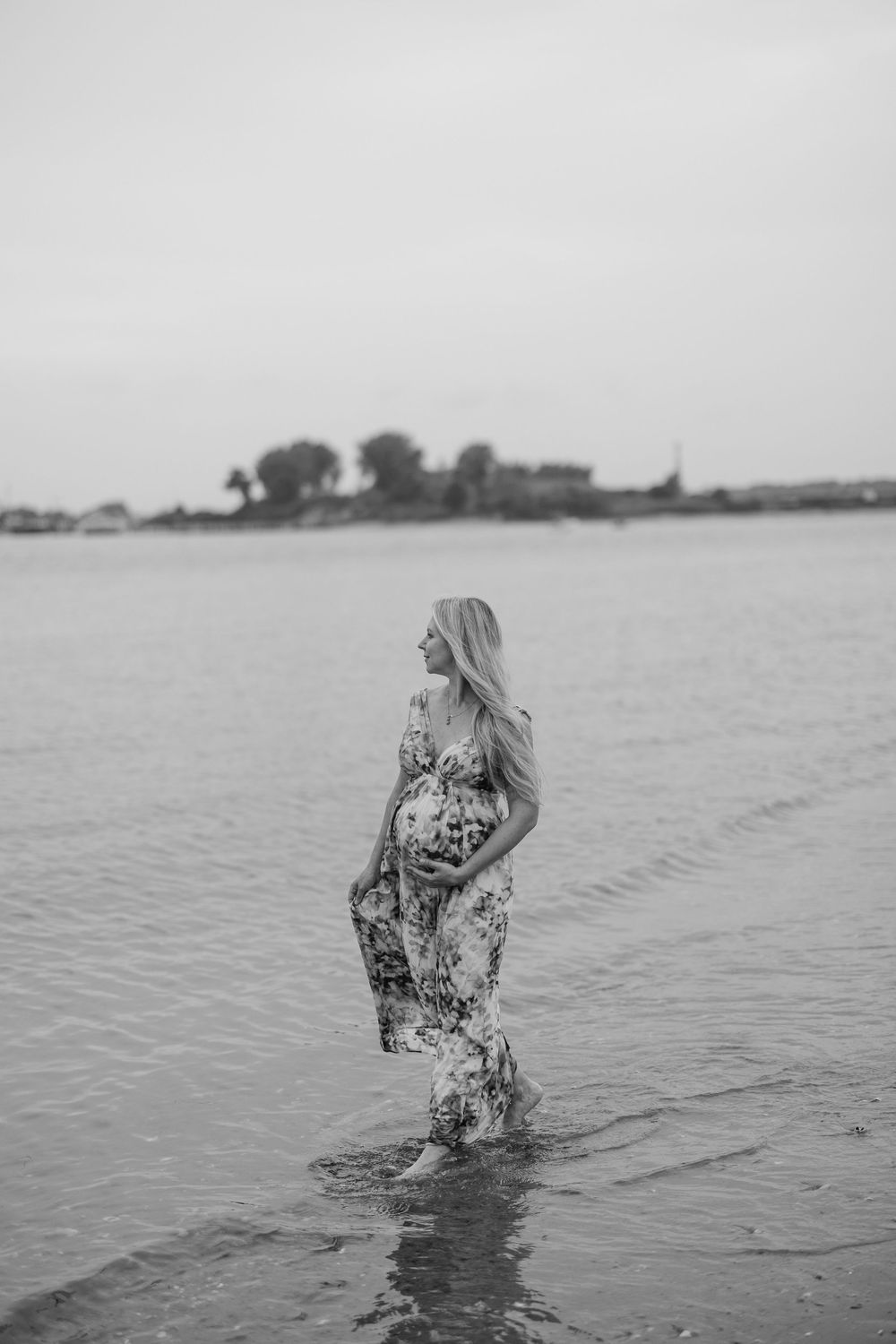 Black and white photo of a pregnant woman in a floral dress standing in shallow water near a shoreline.